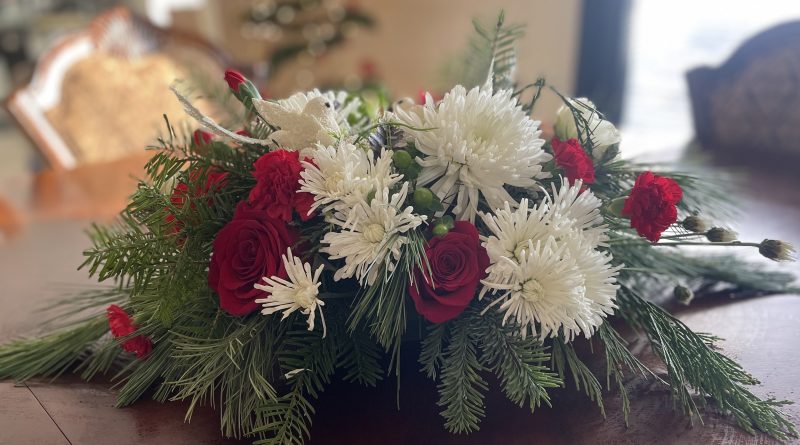 Holiday floral centerpiece with red roses, white mums, evergreen greenery, and silver accents displayed on a dining table